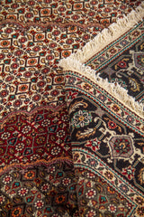 Detalle de alfombra doblada mostrando textura, flecos y diseño central en tonos rojo oscuro, blanco suave, negro y gris