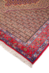 Detalle de esquina de alfombra oriental con campo marrón de pequeños motivos repetitivos, borde rojo y azul, y flecos claros