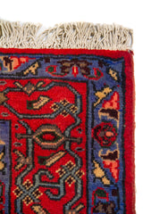 Detalle lateral de alfombra rectangular con guarda decorativa roja y azul, motivos geométricos y terminación con flecos beige