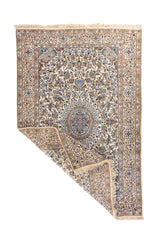 Alfombra oriental en tonos blanco suave y azul con una esquina doblada, mostrando el reverso del tejido y los flecos en uno de sus extremos.