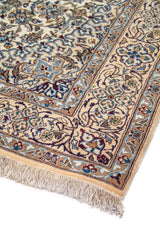 Detalle de esquina de alfombra clásica con motivos florales en azul, celeste y beige sobre fondo blanco suave, terminada con flecos claros.