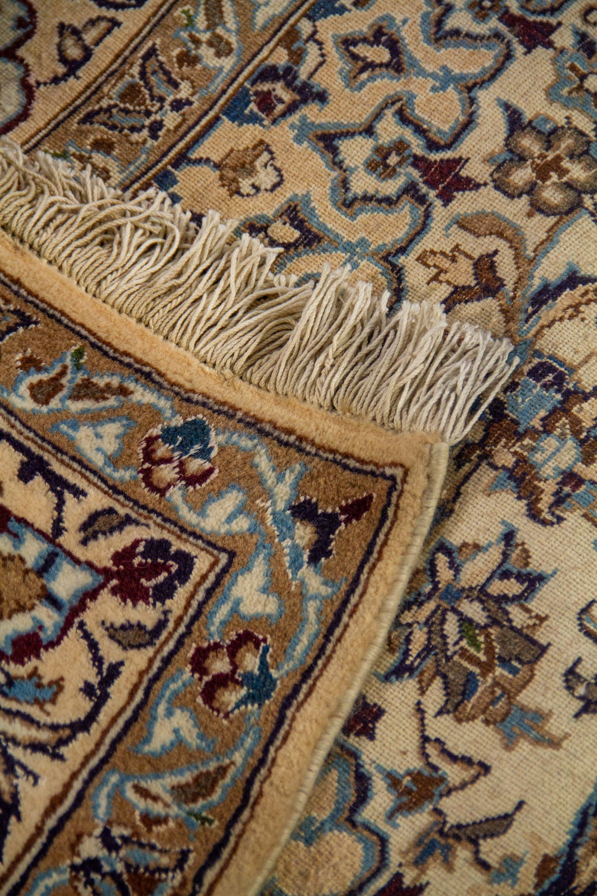 Detalle de alfombra doblada mostrando el reverso del tejido, los flecos y el patrón floral fino sobre fondo claro.