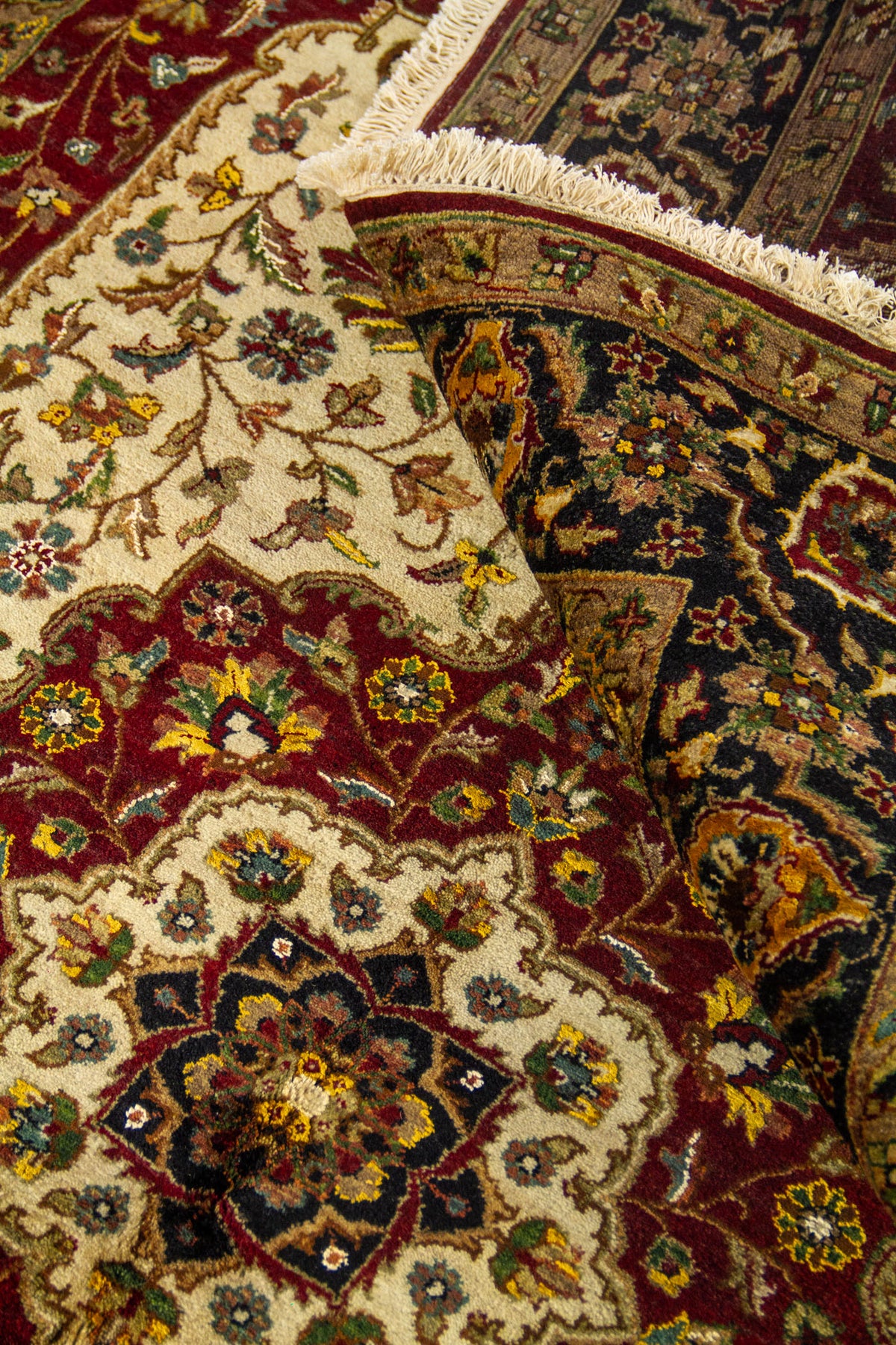 Detalle de alfombra doblada mostrando el reverso del tejido, el borde decorado y los motivos florales en tonos blanco suave, rojo oscuro y negro.