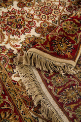 Detalle de alfombra doblada mostrando la textura del tejido, los flecos y los patrones florales en rojo, beige y marrón.