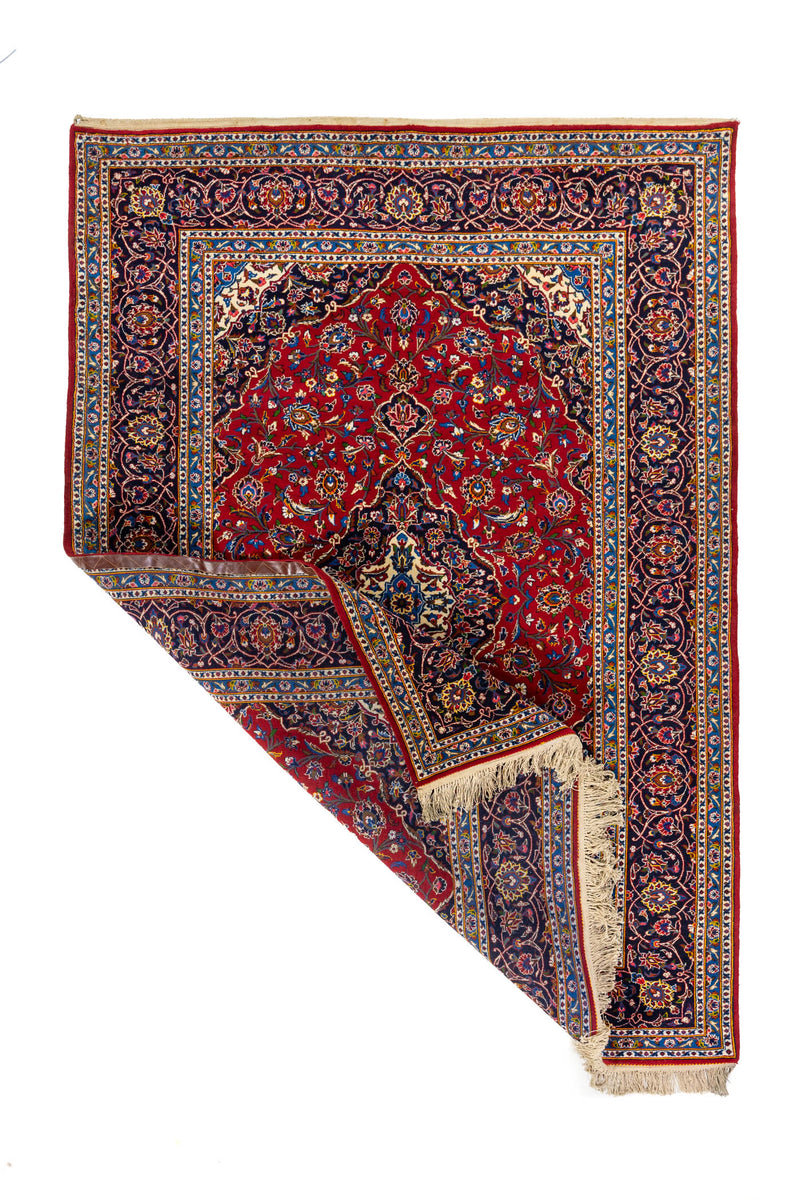 Alfombra clásica rectangular con una esquina doblada que muestra el reverso del tejido, los flecos y el detalle del borde ornamental