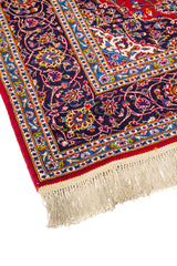 Detalle de esquina de alfombra oriental con fondo rojo, guarda azul floral y flecos claros en uno de los extremos