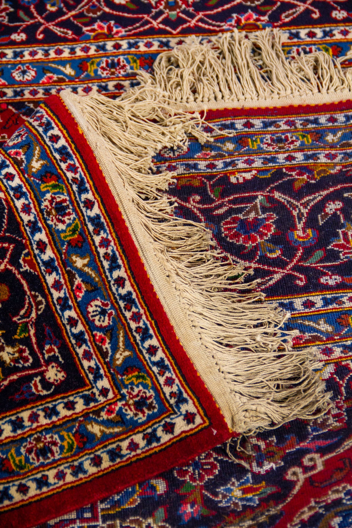 Detalle de alfombra doblada mostrando textura, reverso del tejido y diseño floral ornamental en tonos rojo, azul y beige