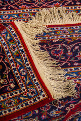 Detalle de alfombra doblada mostrando textura, reverso del tejido y diseño floral ornamental en tonos rojo, azul y beige