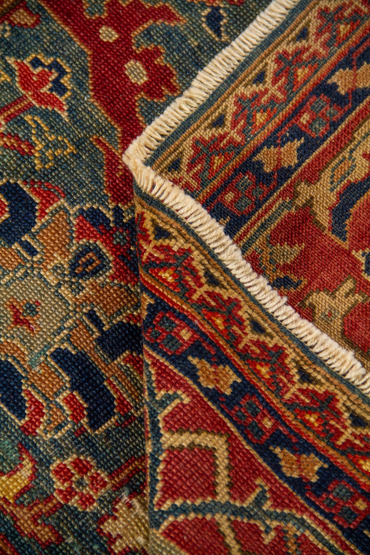 Primer plano de una zona doblada de la alfombra donde se aprecia con claridad la textura del tejido, los flecos y la composición del borde. Los tonos rojo, beige, verde y azul oscuro crean un contraste elegante y lleno de carácter.