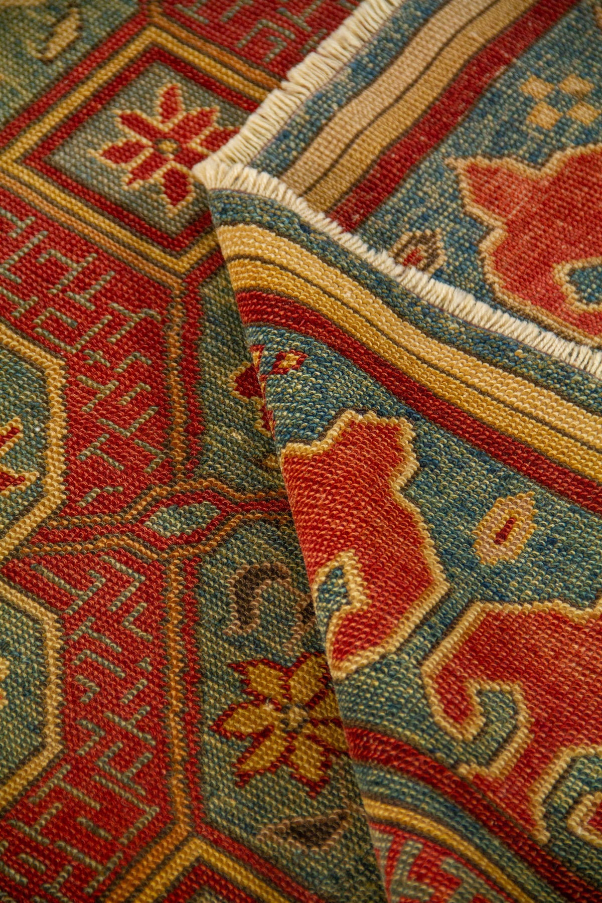 La alfombra aparece parcialmente doblada, mostrando su textura, la definición del tejido y el acabado de los flecos. Esta toma destaca la minuciosidad del diseño y la riqueza ornamental del patrón