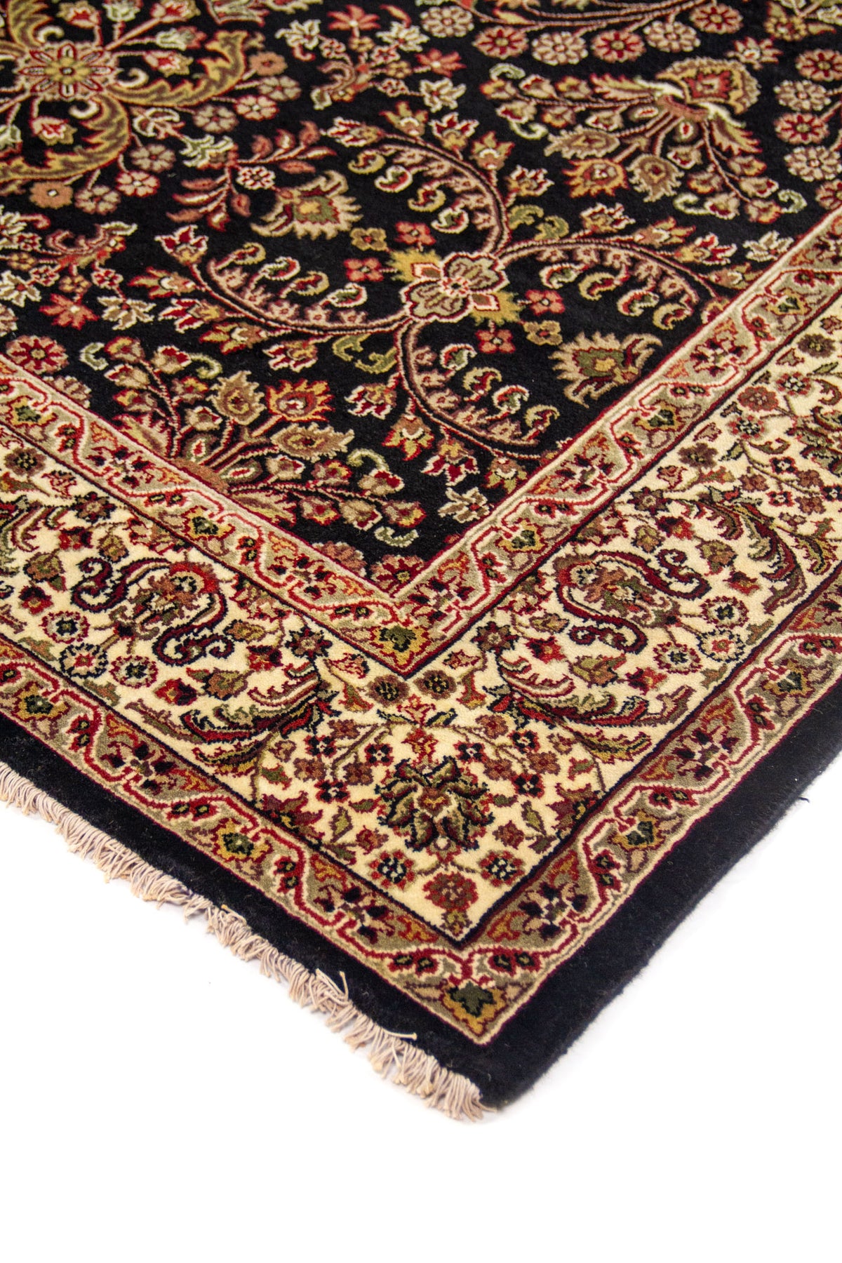 Detalle en perspectiva de esquina de alfombra clásica con fondo negro, motivos florales en rojo, beige y verde, y flecos claros en el borde.