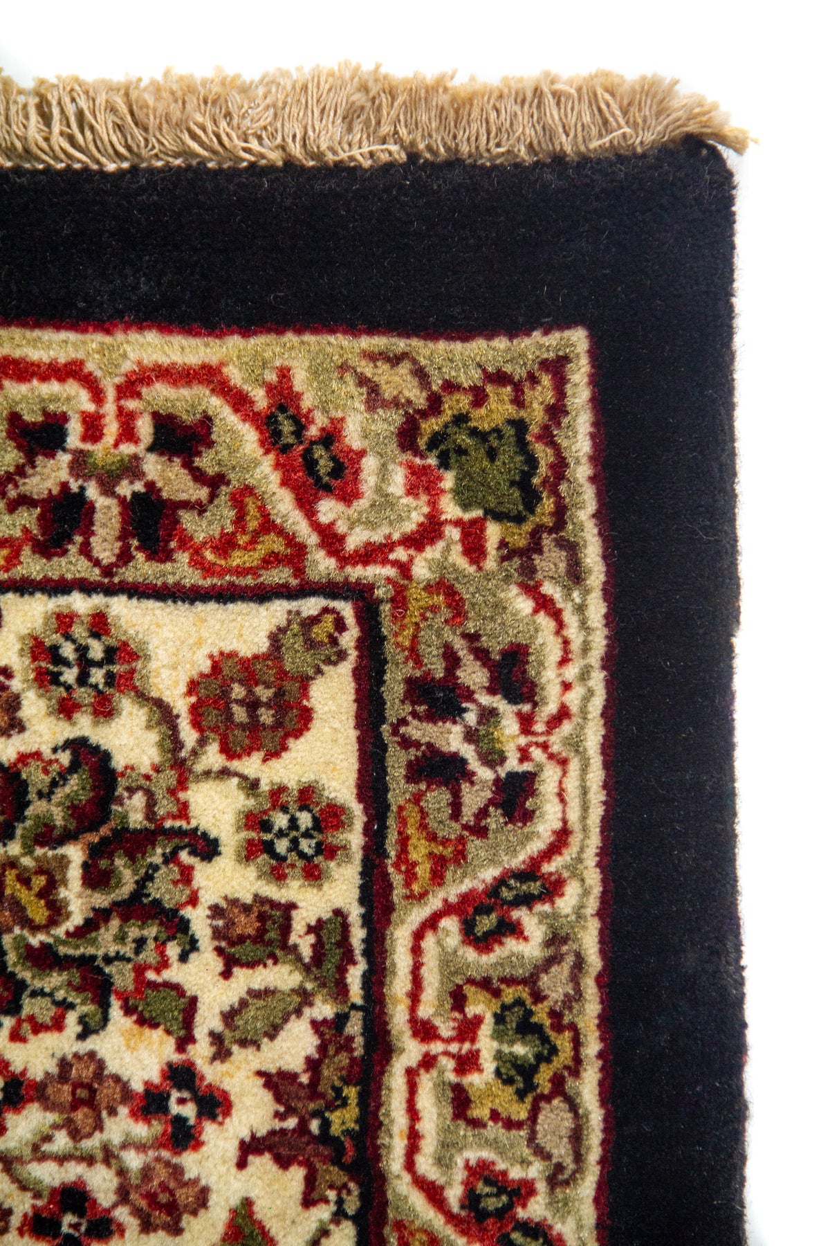 Detalle de esquina de alfombra con marco negro liso, guarda floral blanco suave y flecos beige en uno de sus extremos.