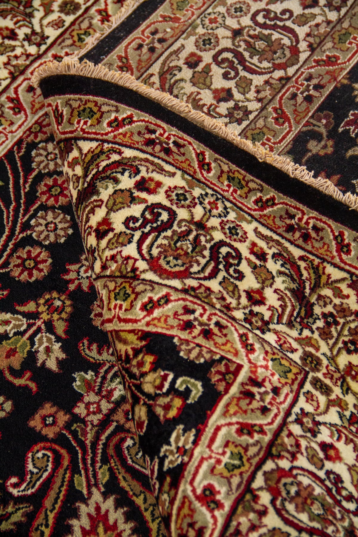 Detalle de alfombra doblada mostrando el reverso del tejido, el borde decorado y los motivos florales sobre fondo oscuro.