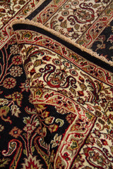Detalle de alfombra doblada mostrando el reverso del tejido, el borde decorado y los motivos florales sobre fondo oscuro.