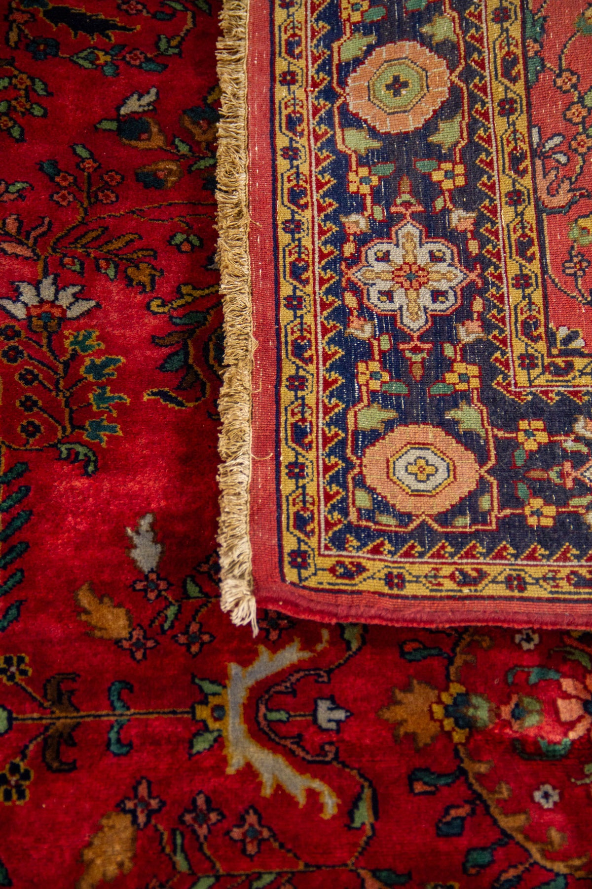 Detalle de alfombra doblada mostrando la textura del tejido, los flecos laterales y el contraste entre el campo rojo y la guarda azul.