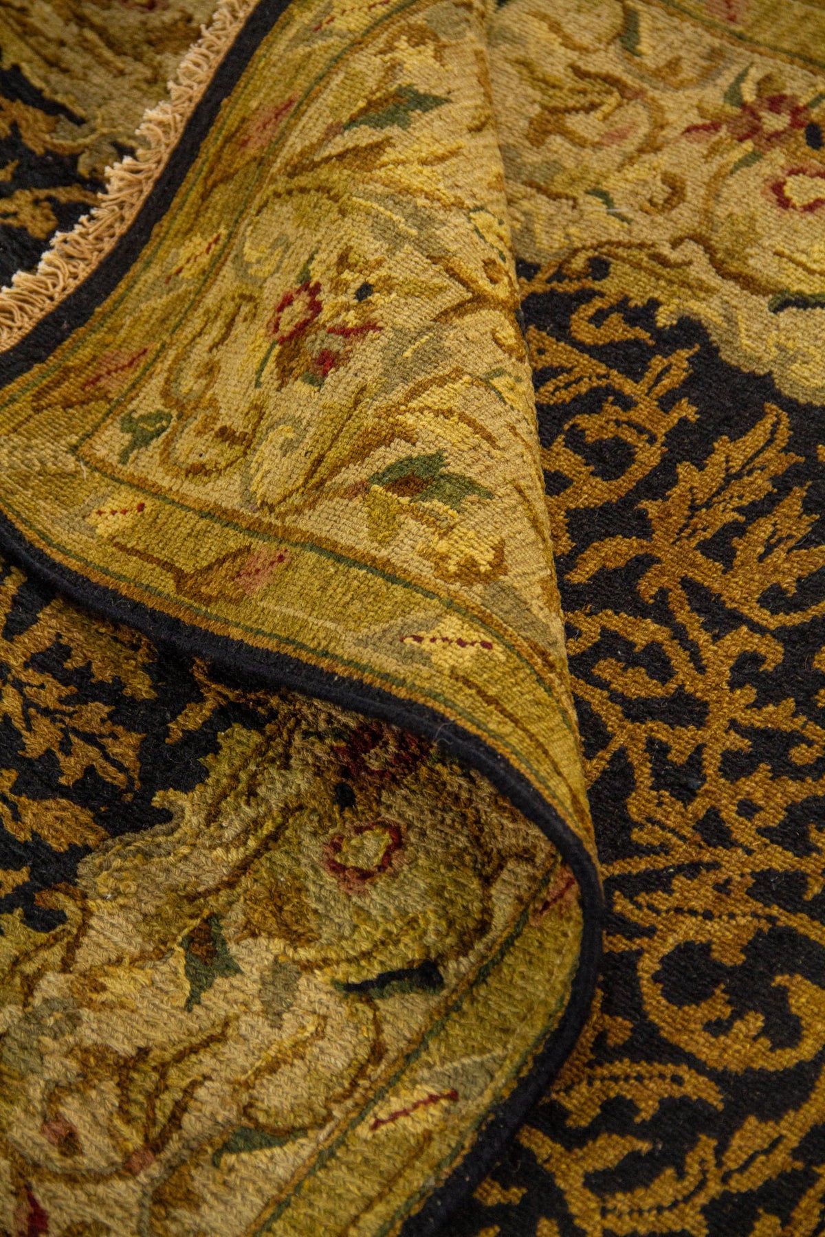 Detalle de alfombra doblada mostrando textura, grosor, acabado de los bordes y diseño ornamental en tonos negro, dorado y beige