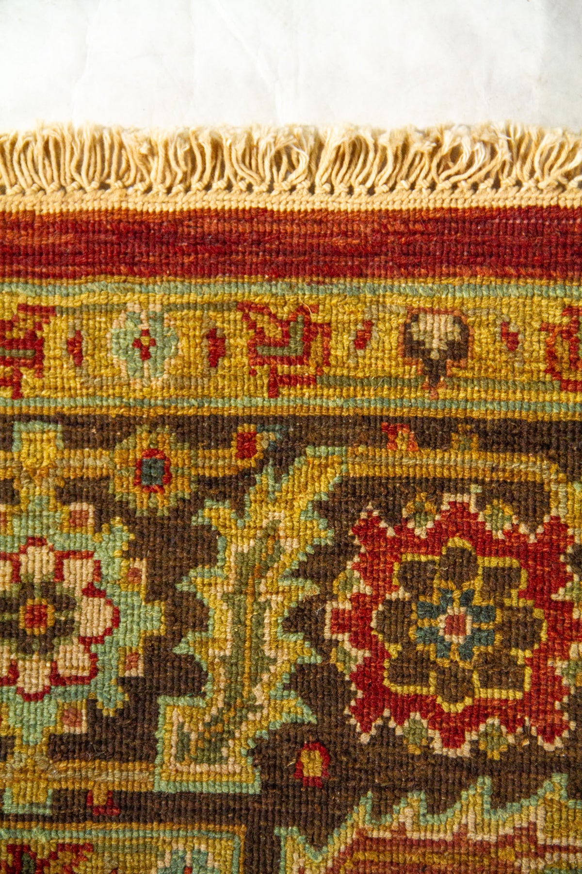 Detalle del borde superior de alfombra oriental con flecos claros y motivos florales geométricos en rojo, marrón, beige y azul.