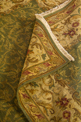 Detalle de alfombra doblada que muestra la textura del tejido, los flecos y el borde floral en tonos verde, beige y dorado