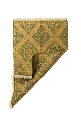 Alfombra verde con esquina doblada que muestra el reverso del tejido y su diseño de rombos decorativos con detalles florales.