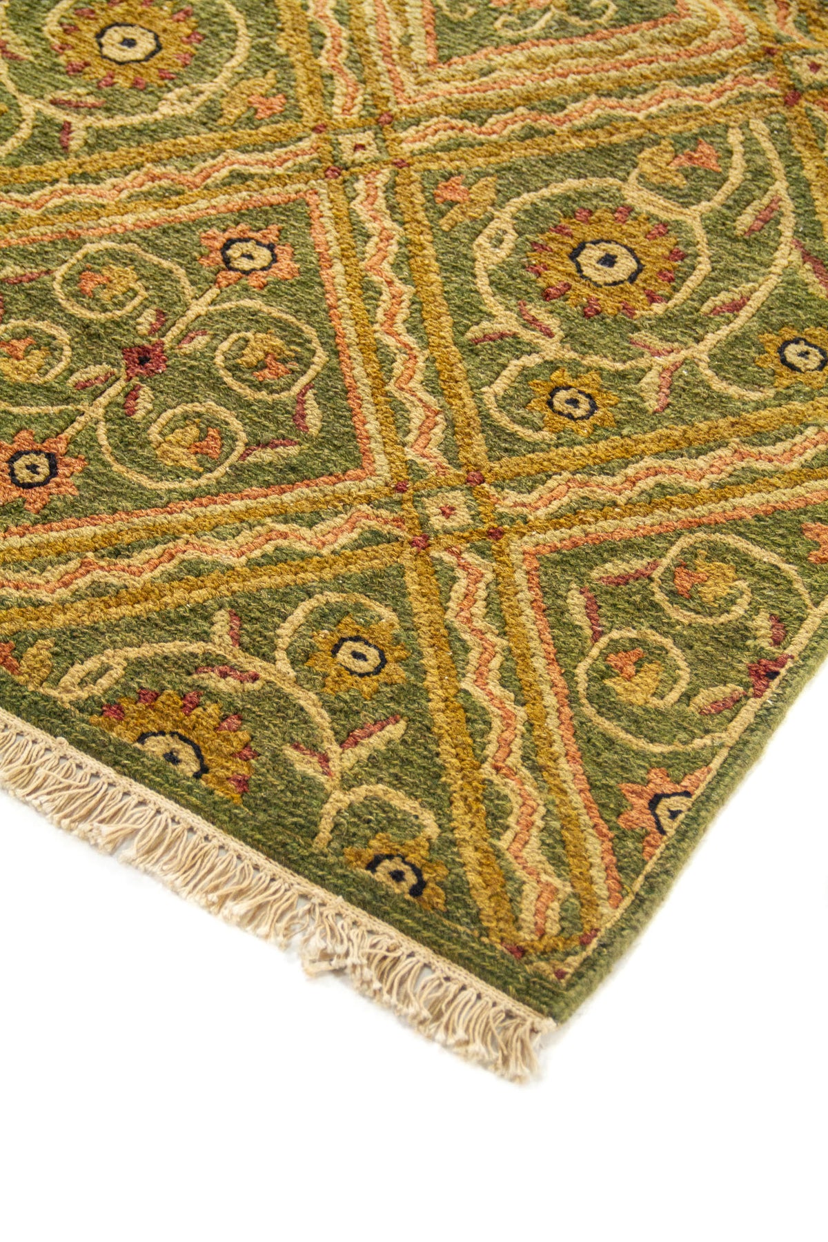 Detalle de esquina de alfombra geométrica en tonos verdes y dorados, con rombos, motivos curvos y flecos color beige.