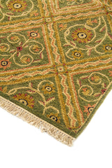 Detalle de esquina de alfombra geométrica en tonos verdes y dorados, con rombos, motivos curvos y flecos color beige.