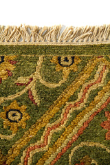 Detalle del borde superior de alfombra verde con flecos claros y motivos geométricos y florales tejidos en tonos cálidos.