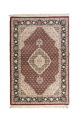 Alfombra clásica en tono rojo con medallón central blanco suave y gris, fondo de pequeños motivos geométricos y guarda floral negra con flecos.
