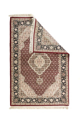 Alfombra clásica rojo doblada en una esquina, mostrando el diseño frontal ornamental y parte del reverso con flecos claros.