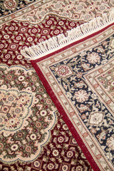 Detalle de alfombra doblada que muestra la textura, el reverso del tejido, los flecos y la combinación de bordes oscuros con centro rojo.
