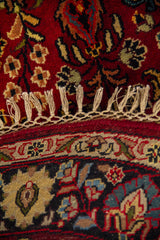 Detalle de alfombra redonda con fondo rojo, diseño floral y flecos artesanales en tono beige.