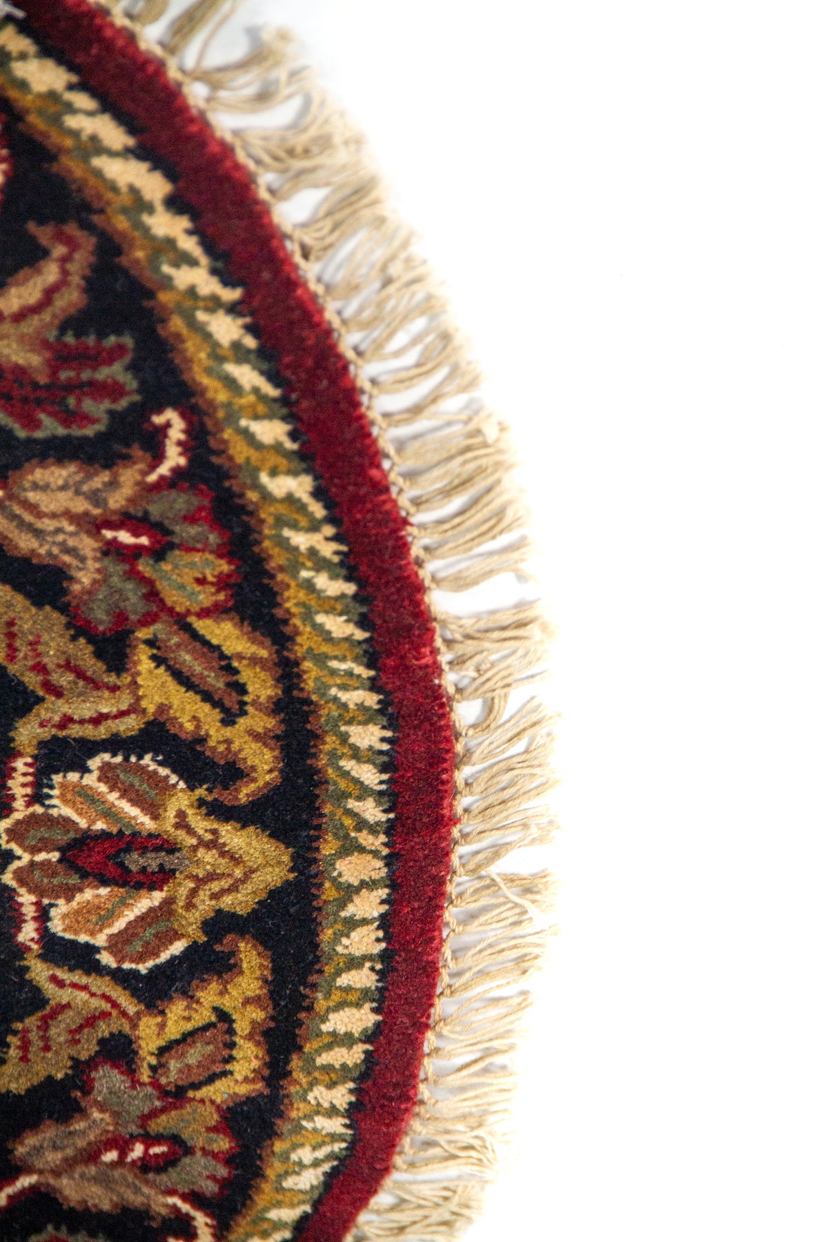 Detalle lateral de alfombra redonda con fondo rojo, borde oscuro ornamental y acabado con flecos.