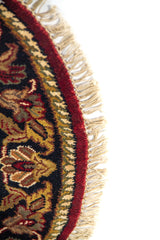 Detalle lateral de alfombra redonda con fondo rojo, borde oscuro ornamental y acabado con flecos.