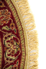 Detalle lateral de alfombra redonda con motivos florales clásicos, borde beige y flecos decorativos.