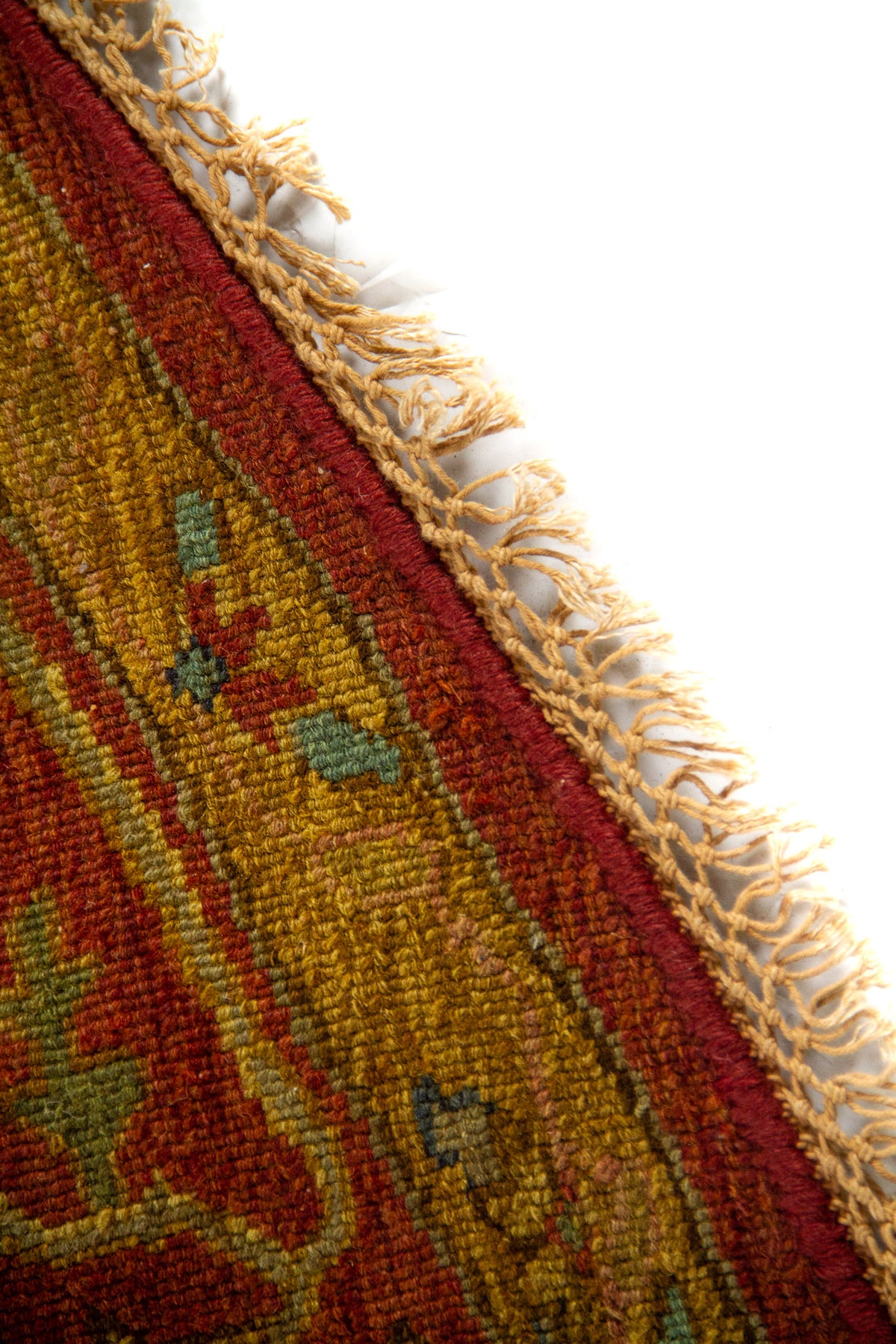 Primer plano del borde de la alfombra con flecos decorativos y tejido en tonos rojo, dorado y verde suave.