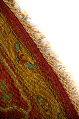 Primer plano del borde de la alfombra con flecos decorativos y tejido en tonos rojo, dorado y verde suave.