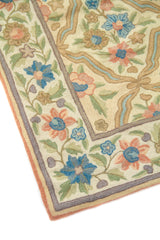 Detalle de esquina de alfombra con guarda floral y patrón interior geométrico curvilíneo, en colores blanco suave, celeste, anaranjado suave, verde y beige
