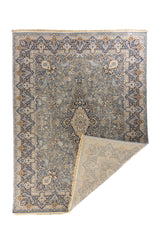 Alfombra gris y azul con una esquina doblada mostrando el reverso y los flecos en detalle.