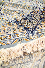 Textura en primer plano de la alfombra doblada, destacando los detalles florales en azul, beige y dorado.