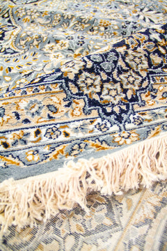 Textura en primer plano de la alfombra doblada, destacando los detalles florales en azul, beige y dorado.