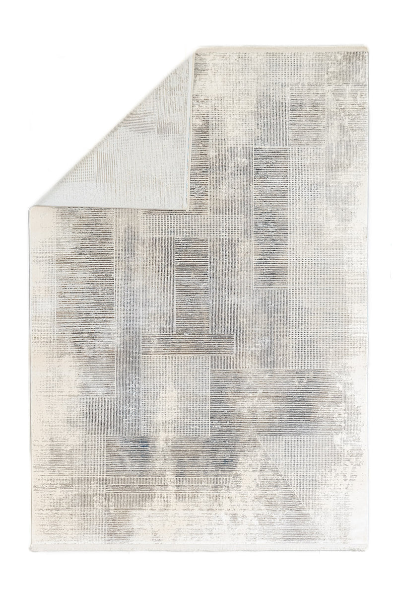 Alfombra con una esquina doblada mostrando el reverso y su patrón geométrico en tonos grises y crema.