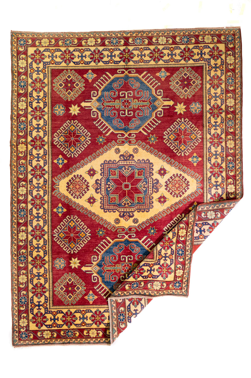 Alfombra Caucasica Kazak rojo y beige de 2.77 x 2.08 mts, vista completa con una esquina doblada; se observan medallones y motivos geométricos caucásicos de fuerte contraste