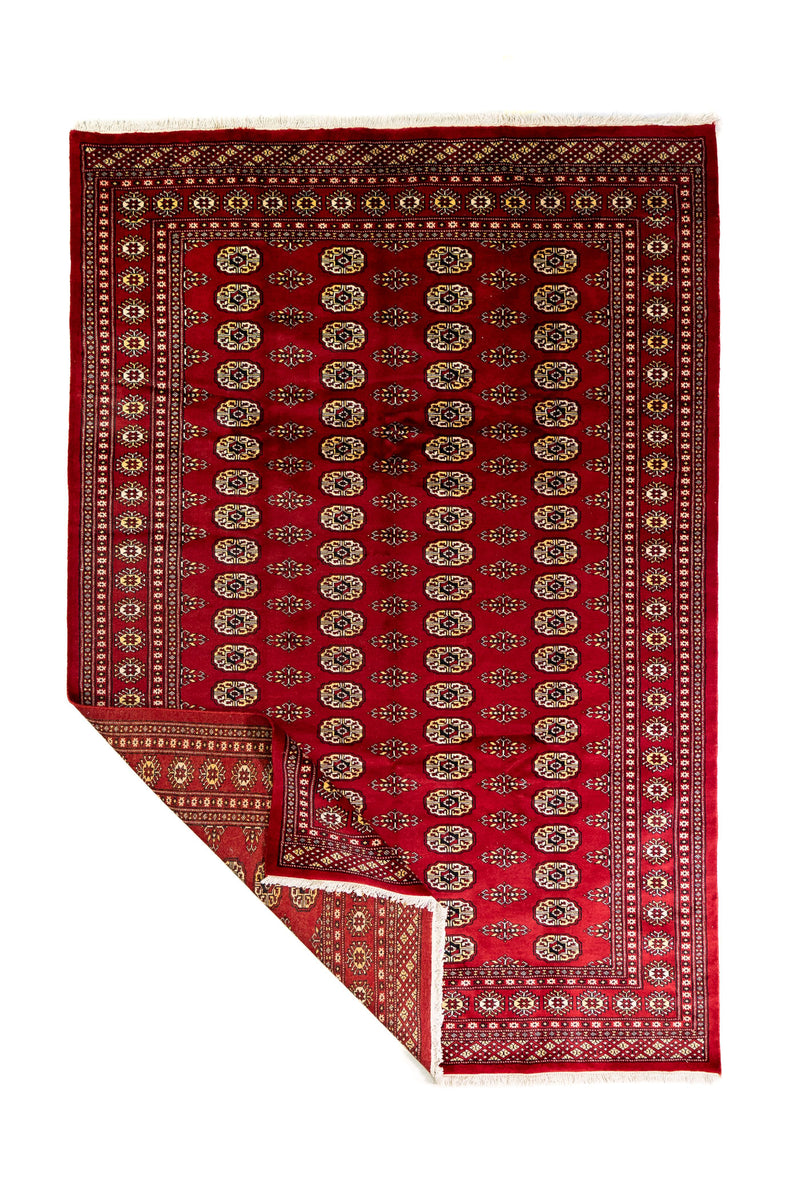 Alfombra Persa Bukara rojo de 1.85 x 2.75 mts, vista completa con una esquina doblada; se observan columnas de medallones ovalados y guardas geométricas