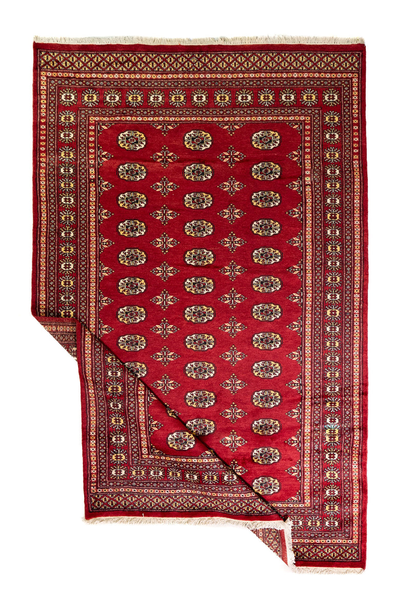 Alfombra Persa Bukara Yamouth rojo de 2.40 x 1.55 mts, vista completa con una esquina doblada; el diseño muestra hileras de medallones ovalados sobre un campo rojo y guardas geométricas densas