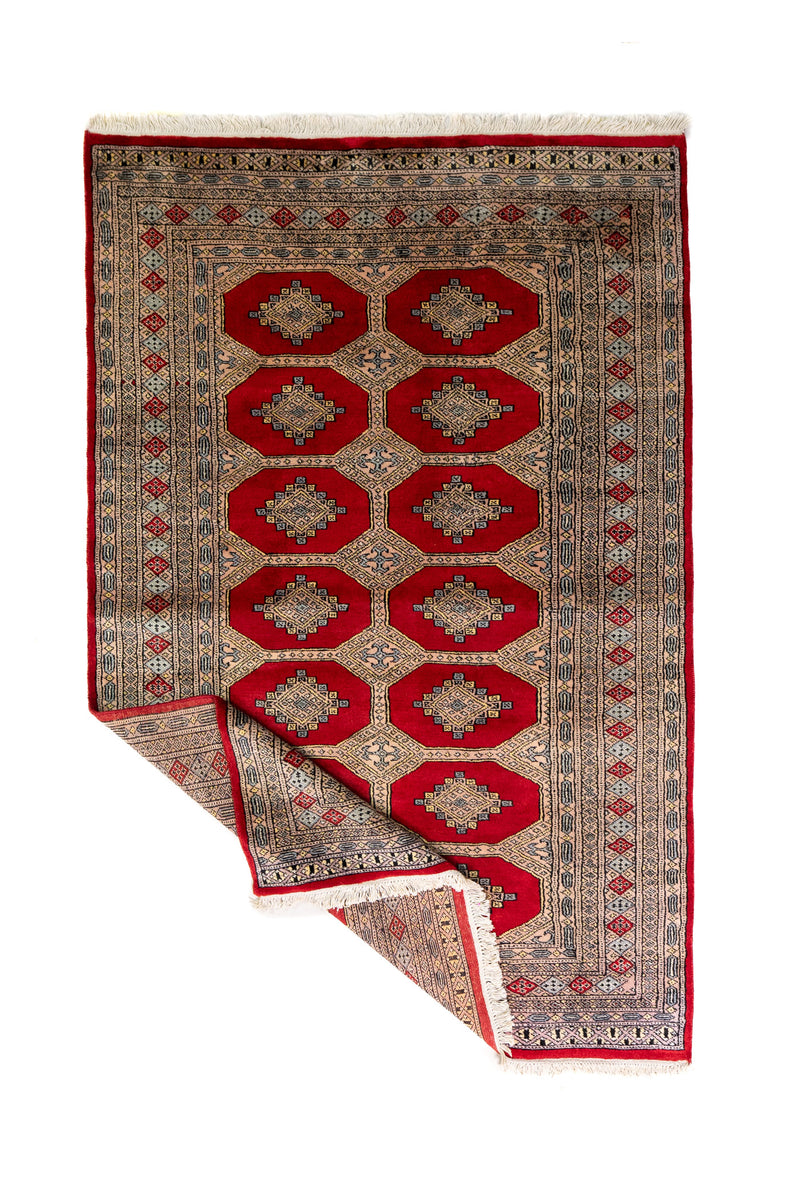 Alfombra Persa Bukara Yamouth rojo de 1.27 x 2.00 mts, vista completa con una esquina doblada; el diseño muestra medallones geométricos repetidos en dos columnas y un marco ornamental de alto contraste