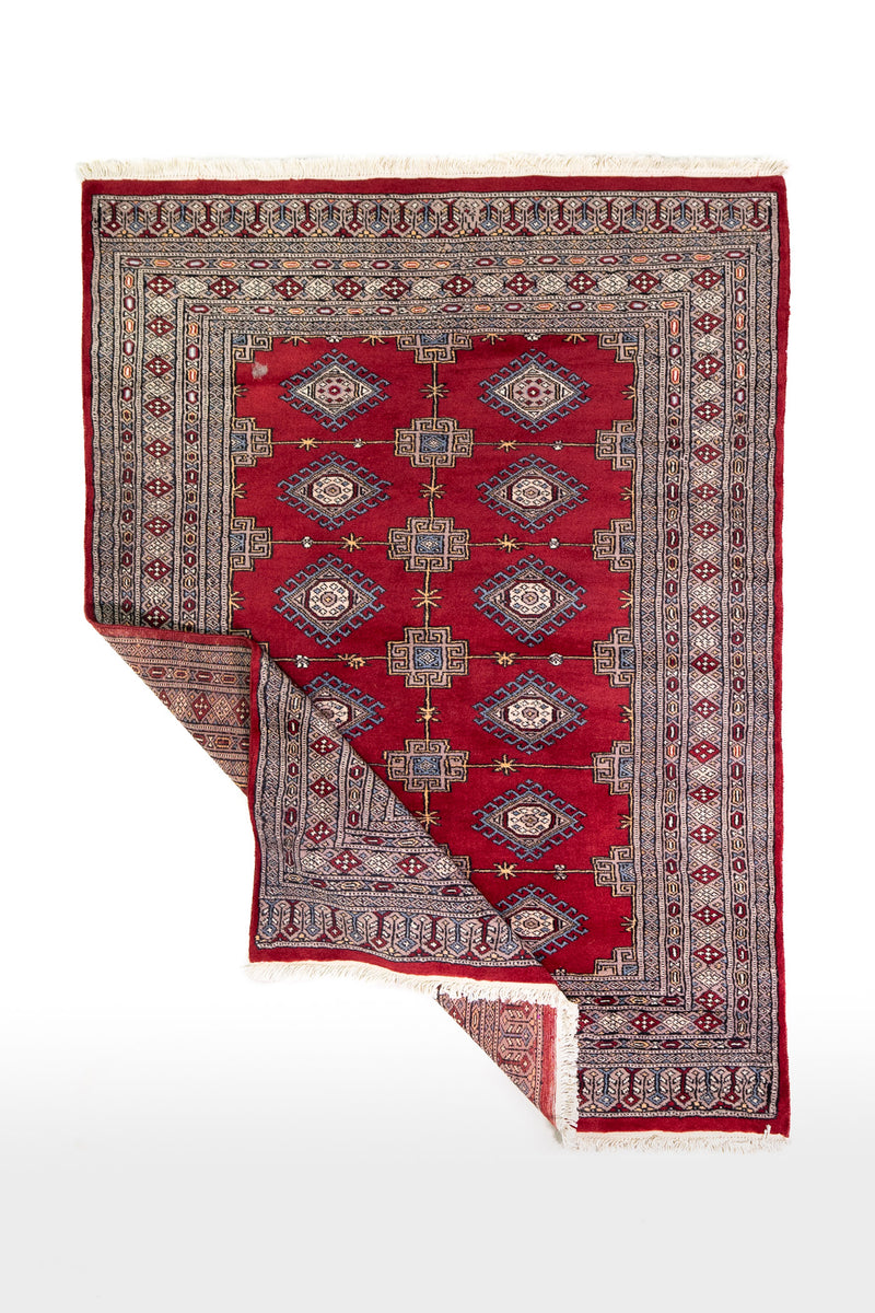 Alfombra Persa Yamouth rojo de 1.24 x 1.68 mts, vista completa con una esquina doblada; el diseño muestra medallones enlazados rojos sobre un campo claro con guardas geométricas azuladas