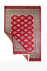 Alfombra Persa Yamouth rojo de 1.27 x 1.88 mts, vista completa con una esquina doblada; el diseño muestra una retícula de rombos rojos enlazados con guardas geométricas de alto contraste