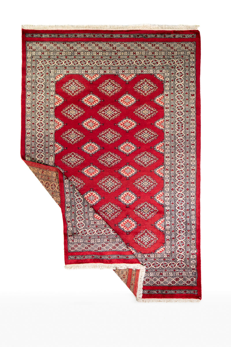Alfombra Persa Yamouth rojo de 1.27 x 1.88 mts, vista completa con una esquina doblada; el diseño muestra una retícula de rombos rojos enlazados con guardas geométricas de alto contraste
