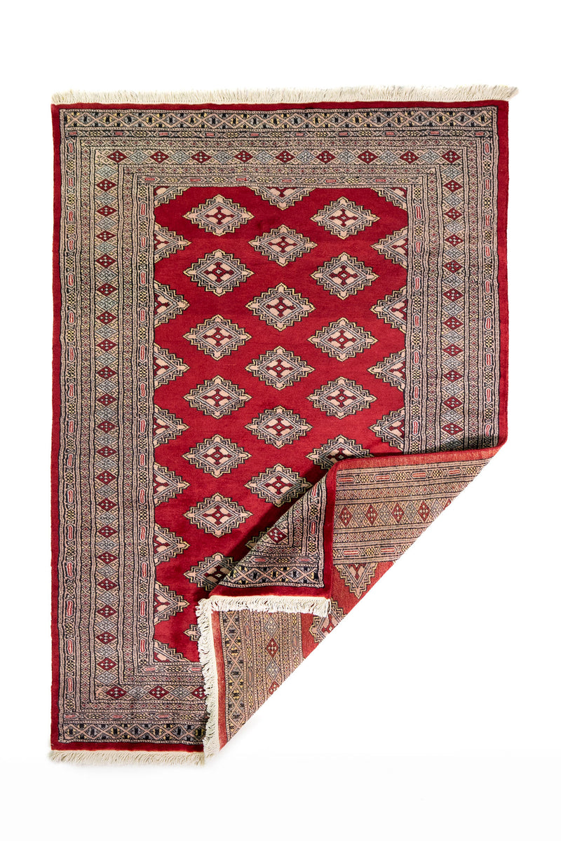 Alfombra Persa Yamouth rojo de 1.24 x 1.78 mts, vista completa con una esquina doblada; el diseño muestra una cadena de rombos rojos enlazados sobre un campo claro y guardas oscuras