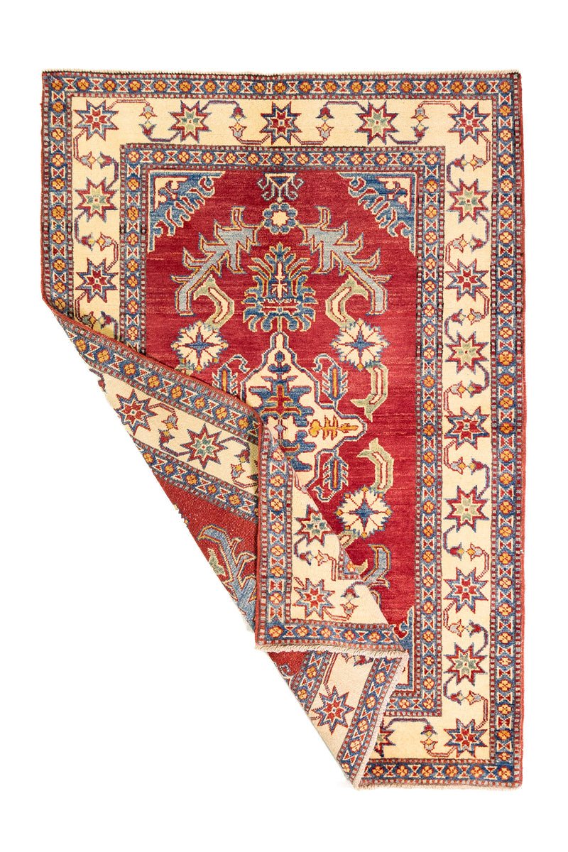 Alfombra Caucasica Kazak bordó y beige de 1.14 x 1.65 mts, vista completa con una esquina doblada; el diseño muestra un gran motivo central de lectura geométrica sobre un campo bordó y guardas beige