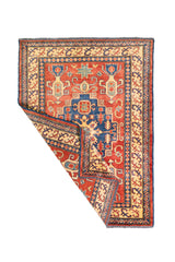 Alfombra Caucasica Kazak ladrillo y beige de 1.07 x 1.45 mts, vista completa con una esquina doblada; el diseño muestra un medallón central azul rodeado por motivos geométricos sobre un campo ladrillo
